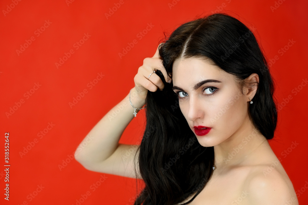 Fototapeta premium The concept of long beautiful hair. Close-up Portrait of a pretty brunette model girl with excellent makeup and luxurious hair on a red background in the studio.