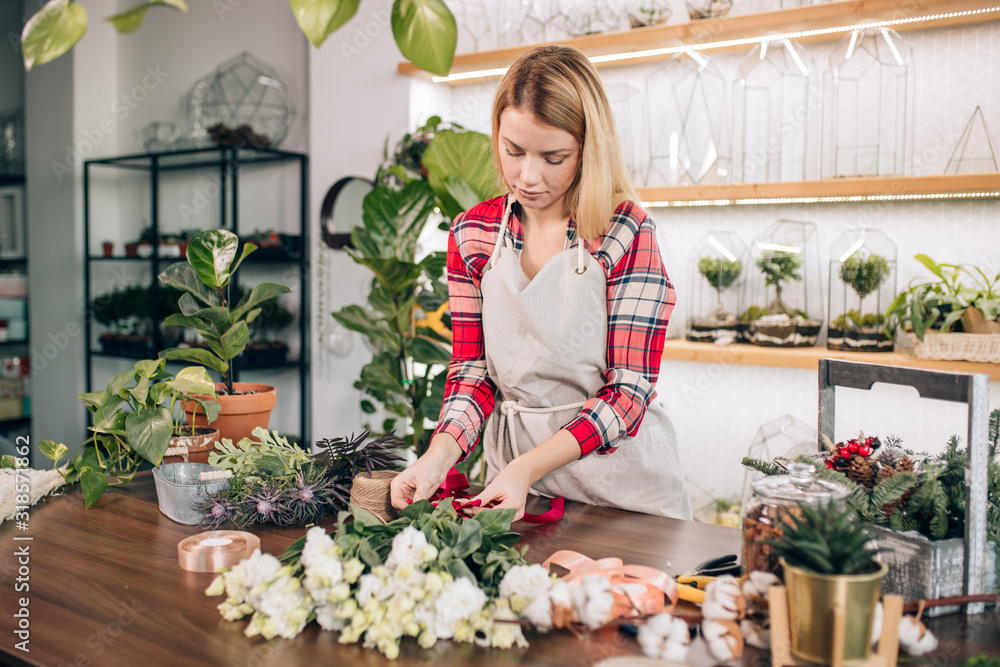 flowers composing and decoration by young lady with blonde hair, wearing red checkered shirt, isolated in light room full of green plants. botany, flowers concept
