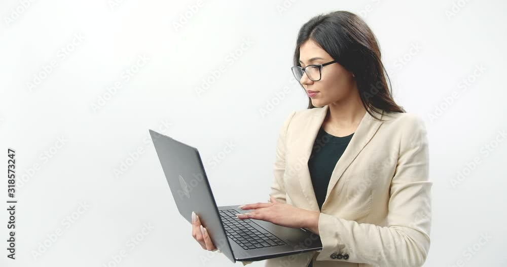 Side view of serious business woman in eyeglasses and formal clothing holding laptop and using it while standing in studio with white background. Concept of modern technology Side view of serious business woman in eyeglasses and formal clothing holding laptop and using it while standing in studio with white background. Concept of modern technology