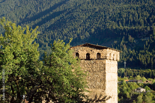 The top of the Svan tower, stone, quadrangular, against the backdrop of a mountain covered with green dense forest. Windows are visible under the roof of the tower. Sunny day. Mestia, Svaneti, Georgia