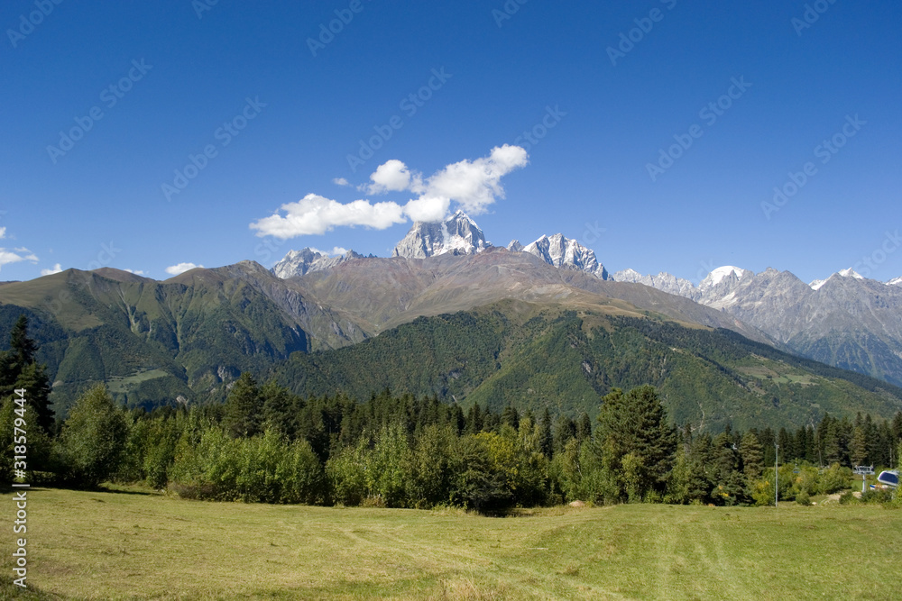 Mountain landscape. In the background is the top of Mount Ushba, partially covered by small clouds. In the foreground are green trees and a field. Bright autumn day in Svaneti, Georgia