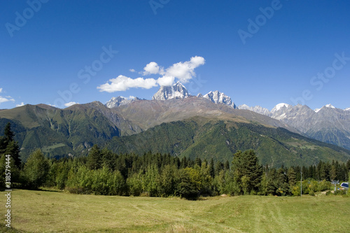 Mountain landscape. In the background is the top of Mount Ushba, partially covered by small clouds. In the foreground are green trees and a field. Bright autumn day in Svaneti, Georgia