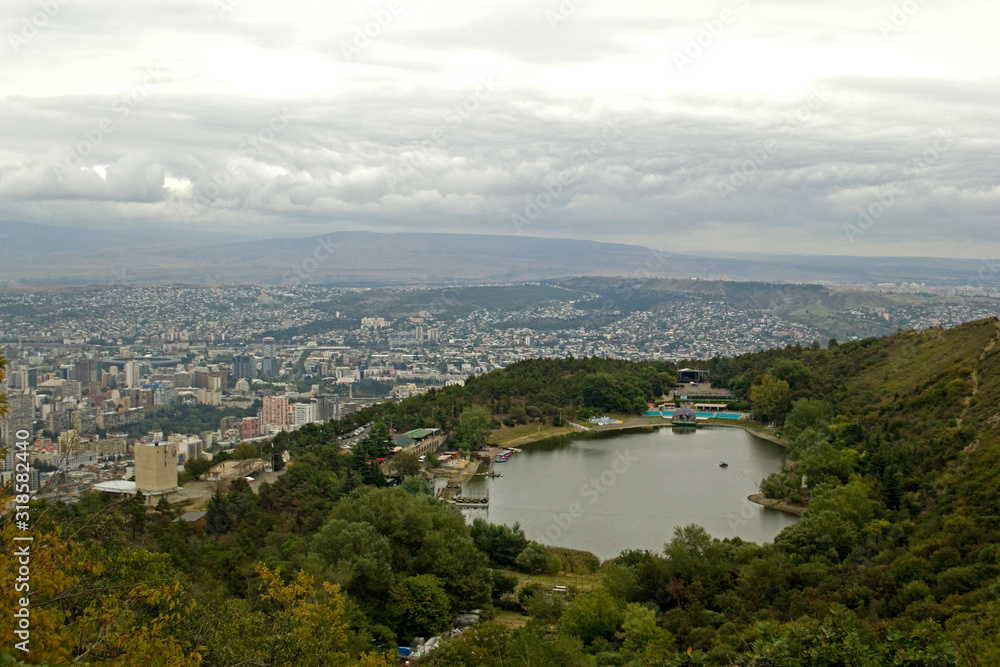 Turtle lake. A small lake on the slopes of Mtatsminda, surrounded by trees along the banks. In background is the capital of Georgia, the city of Tbilisi. On the horizon - mountains and gray cloudy sky