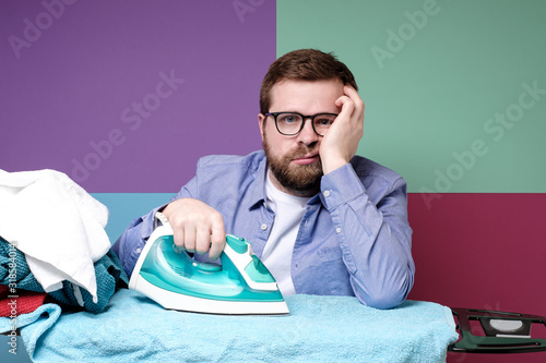 Obraz na plátně Unhappy bearded man with glasses is ironing clean linen on the board, he is tired of housework and looks sadly at the camera