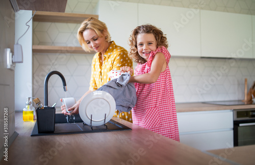 A cute small girl with mother indoors in kitchen at home, washing up dishes.