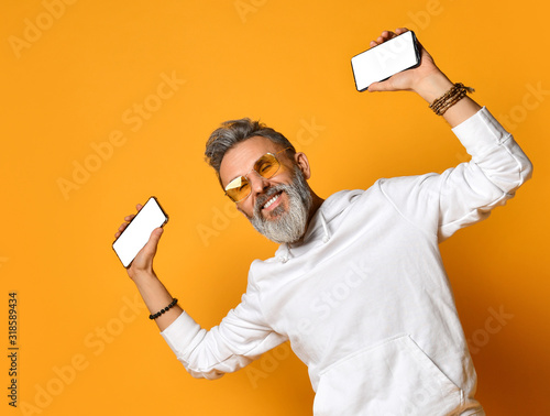 Stylish smiling grey-haired hipster adult man in white hoodie and sunglasses standing holding smartphones in hands photo