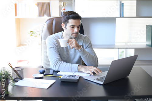 A young man working at a desk with a laptop. Calculator, notepad, pencil, pen and cup of coffee on the side.