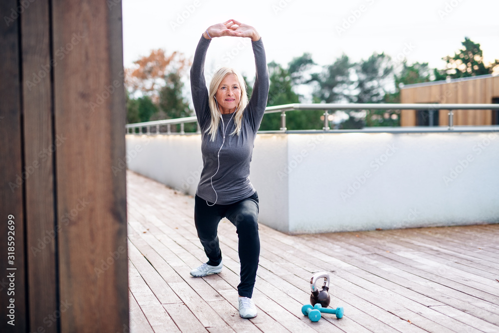 A senior woman doing exercise outdoors, stretching. - obrazy, fototapety, plakaty 
