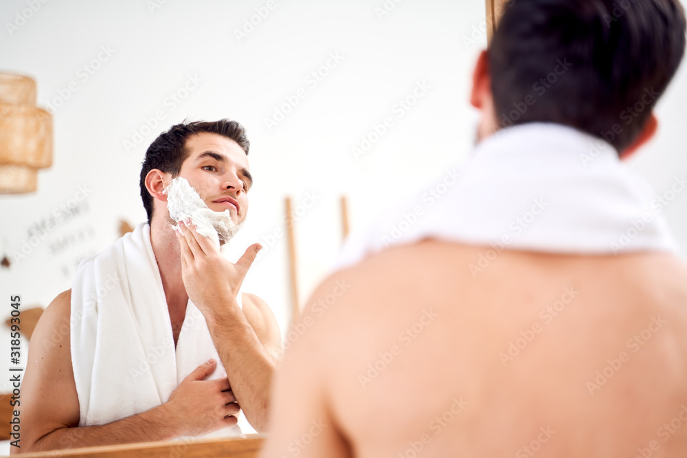 Brunet man with towel on shoulders spreads face with shaving foam while standing in bath near mirror
