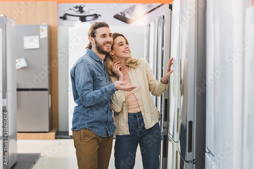 smiling boyfriend pointing with hand and girlfriend touching fridge in home appliance store