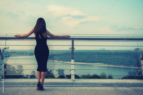 Fashion model wearing black elegant dress posing outdoor at river Dnirpo bank in Kyiv, Ukraine. Young beautiful brunette caucasian woman walking summer streets. Beautiful girl, urban portrait. photo