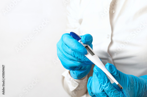 The doctors hands in blue gloves hold a electronic thermometer on light background, copy space. Checking the patients high temperature, symptoms of a virus, infection or illness. Diagnosis of health.