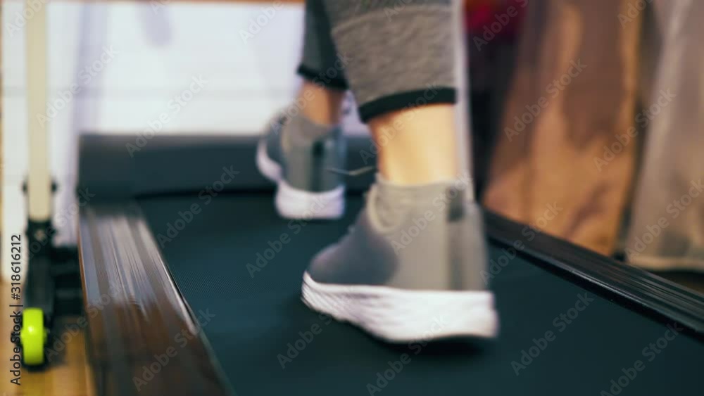 CU: young lady in grey sneakers walks along modern running machine in light room close backside low angle shot CU: young lady in grey sneakers walks along modern running machine in light room close backside low angle shot