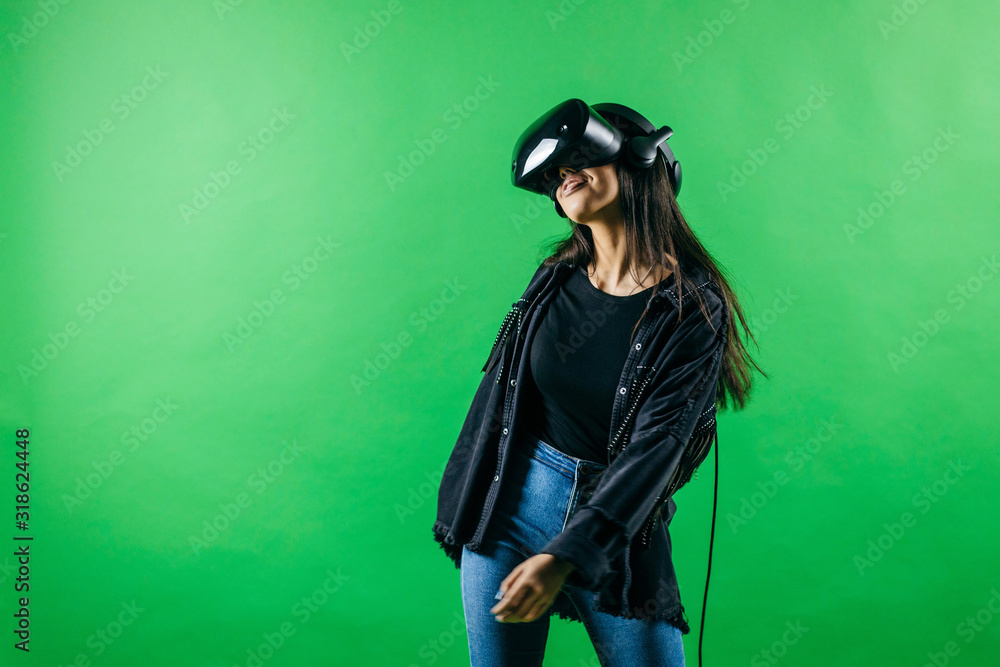 Mod curly dark haired girl dressed in black denim jacket uses the virtual reality glasses on her head and gloves in hands in the studio over green background Mod curly dark haired girl dressed in black denim jacket uses the virtual reality glasses on her head and gloves in hands in the studio over green background