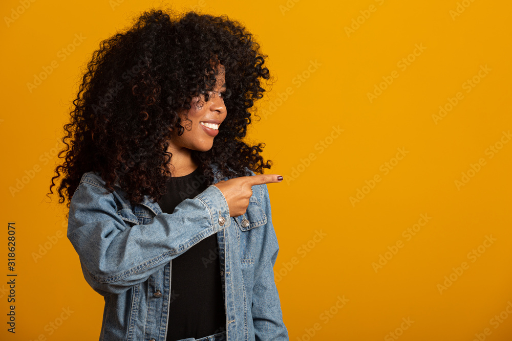 Cheerful Afro woman points away on copy space, discusses amazing promo, gives way or direction, wears yellow warm sweater, has pleasant smile, feels optimistic, isolated over yellow background. Cheerful Afro woman points away on copy space, discusses amazing promo, gives way or direction, wears yellow warm sweater, has pleasant smile, feels optimistic, isolated over yellow background.