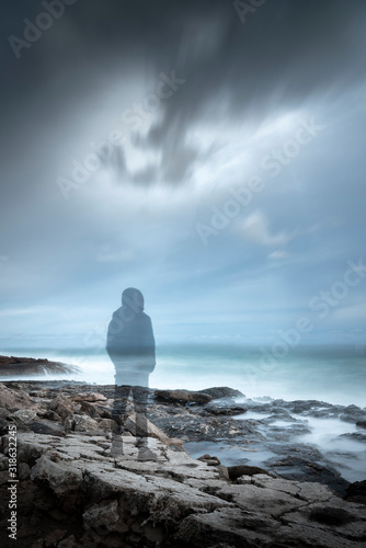 Transparent gloomy scene of a hooded man like a ghost, dressed in black in a cove, looking at the maritime horizon. Inspirational and emotional aesthetics.
