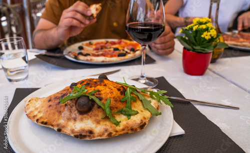 Baked calzone on the plate. Closed type of pizza, decorated with basil twigs on a restaurant table background.