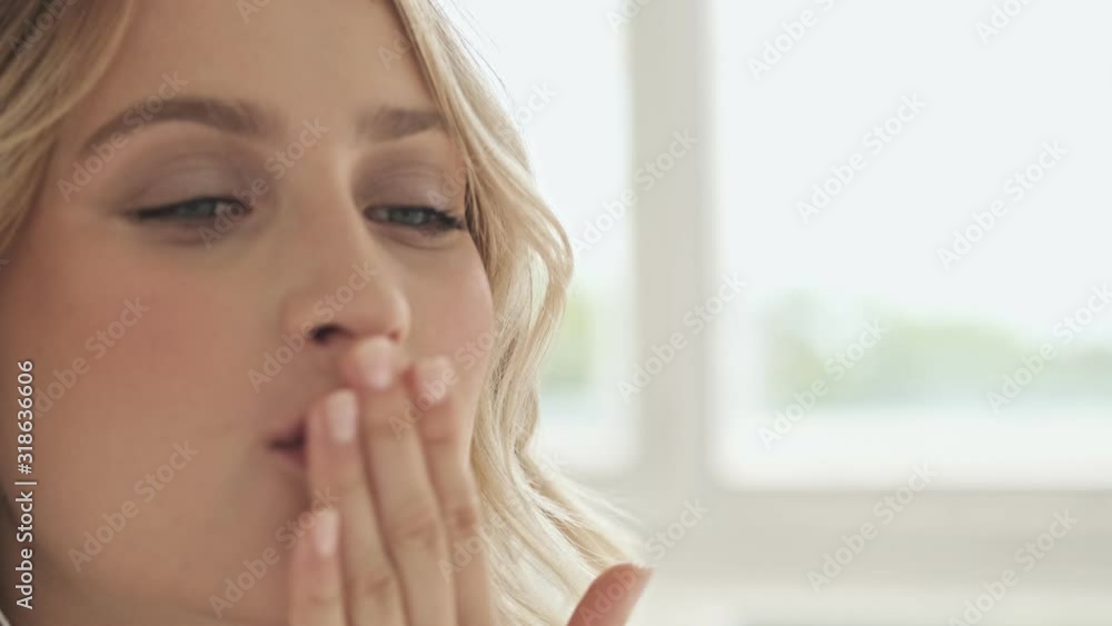Close up view of an attractive smiling woman with blond hair is sending air kisses while having a video call on her cellphone with wired headphones isolated over fabric background Close up view of an attractive smiling woman with blond hair is sending air kisses while having a video call on her cellphone with wired headphones isolated over fabric background