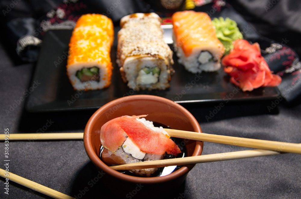 Sushi is a Japanese dish that consists of glutinous rice with fish, seafood and vegetables. On a black background is a plate with sushi, ginger, soy sauce and wasabi, wooden sticks.