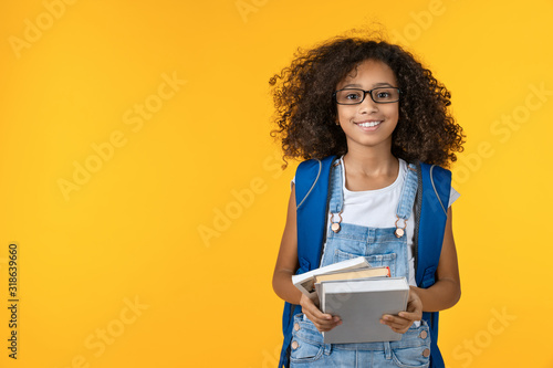 Cheerful young african girl kid in eyeglasses holding notebook and books for study isolated over yellow background photo