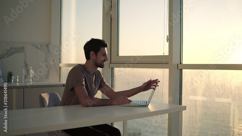 European dark haired man sitting close to panoramic window on studio kitchen - looking on laptop deskop and cheerfully smiling, chatting with friends. Slow motion. Side view photo