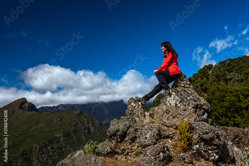 Woman at climbing the rock, sitting on the top with a smile and enjoying view on other steep, green mountain range. Roque de las Muchachos at back hidden in clouds. Rota Volcanica, GR131, 