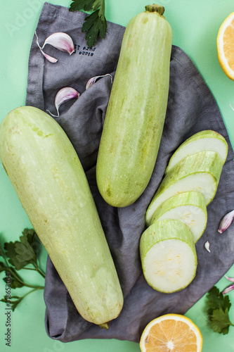 Green fresh ripe zucchini whole and cut into slices on a cloth napkin surrounded by sprigs of parsley, garlic and lemon.