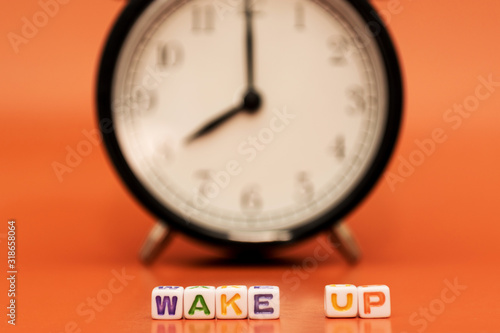 "Wake up" is written by cubes with letters with a black and white classic alarm clock on backside on orange background