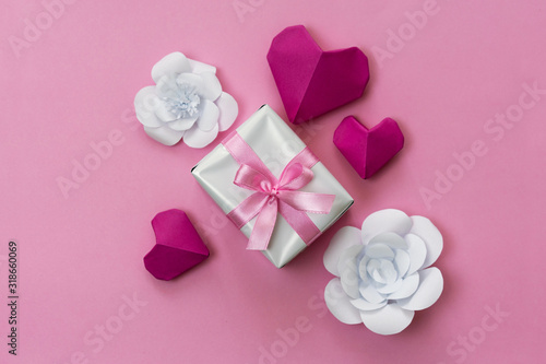 Romantic gift flat lay photo with a box wrapped with pink ribbon, origami hearts and white paper flowers. © juhrozian