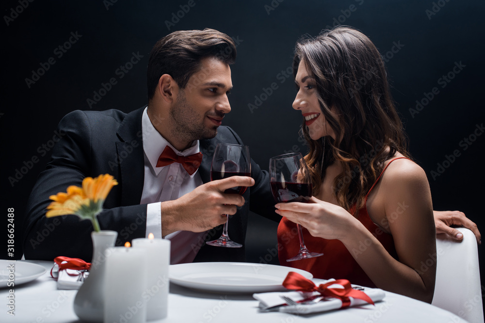 Side view of smiling couple with wine glasses looking at each other at served table isolated on black Side view of smiling couple with wine glasses looking at each other at served table isolated on black