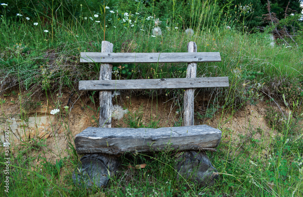 Wooden empty bench in the forest. Rustic bench and fresh green grass near it. Homemade bench made of wooden logs near a forest path. Resting seat while hiking. Wooden empty bench in the forest. Rustic bench and fresh green grass near it. Homemade bench made of wooden logs near a forest path. Resting seat while hiking.