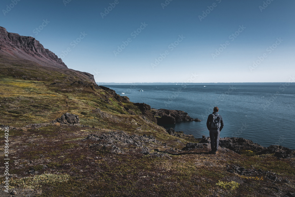 Young man hiker in the arctic landscape of Disko Bay in Greeland in Summer. Blue Sky and green meadows. Arctic Circle Trail with temple mountains. Disko Island and Village of Qeqertarsuaq,