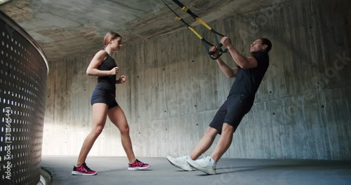 Strong Caucasian female personal trainer explaining pull ups exercise to motivated guy, effort powerful attractive man using holding trx loops outdoor urban concrete dressed in black sportswear