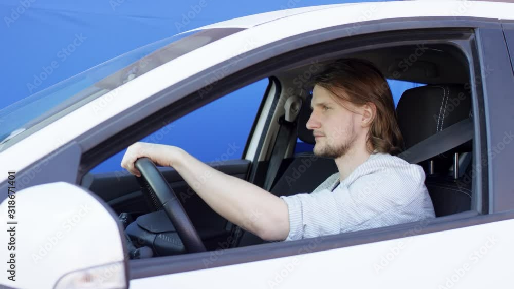 Funny guy is driving a car and taking selfie. Brunette man in T-shirt and jacket is sitting in auto against blue background. Young driver is making funny face and taking photos on mobile phone. Funny guy is driving a car and taking selfie. Brunette man in T-shirt and jacket is sitting in auto against blue background. Young driver is making funny face and taking photos on mobile phone.