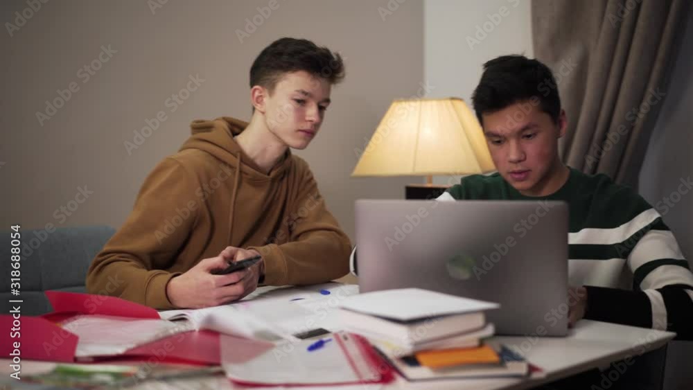 Concentrated teenage Asian boy showing laptop screen to his Caucasian groupmate using smartphone. Two handsome male college students studying online. Distant education, lifestyle, modern technologies. Concentrated teenage Asian boy showing laptop screen to his Caucasian groupmate using smartphone. Two handsome male college students studying online. Distant education, lifestyle, modern technologies.