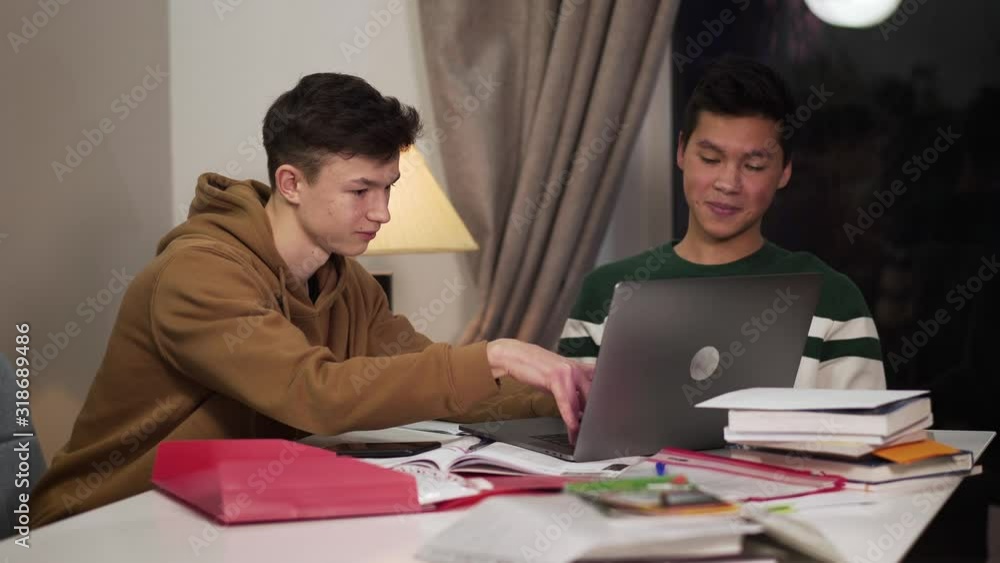 Two intelligent smiling college students sitting at the table and using laptop. Happy Caucasian and Asian teenagers studying online and giving high five. Happiness, intelligence, education. Two intelligent smiling college students sitting at the table and using laptop. Happy Caucasian and Asian teenagers studying online and giving high five. Happiness, intelligence, education.