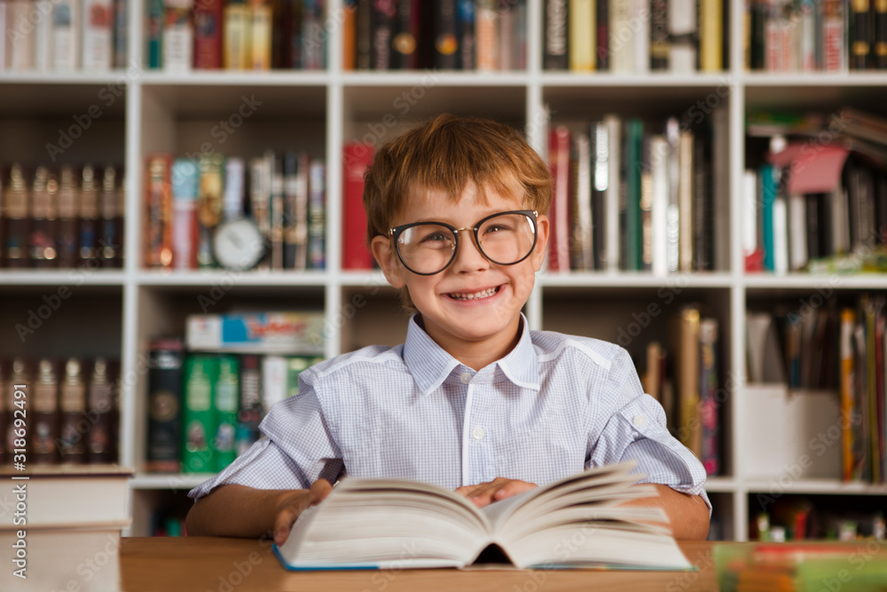 little boy wearing spectacles reading book at table about a bookcase at home. homework little boy wearing spectacles reading book at table about a bookcase at home. homework