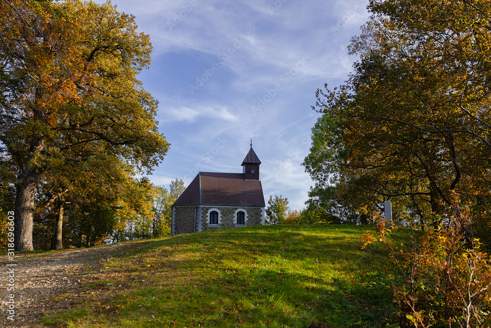 Small chapel in the forest, on top of the hill, with autumn foliage and blue sky with clouds Small chapel in the forest, on top of the hill, with autumn foliage and blue sky with clouds