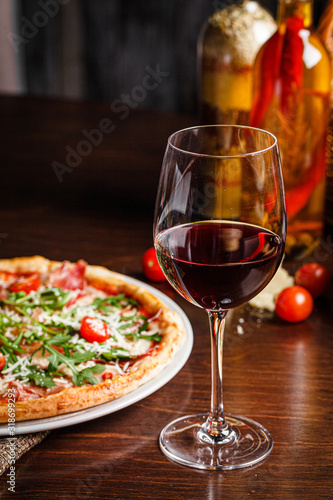 Italian food. Classic thin pizza with large sides, prosciutto, cherry tomatoes, arugula, parmesan cheese. Serving dishes in a restaurant on a white plate with red wine.