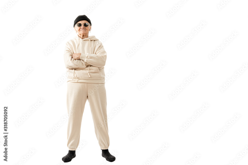 Stylish old hipster woman in beige hoodie black hat and sunglasses stands with crossed hands and looking at the camera isolated over white background Stylish old hipster woman in beige hoodie black hat and sunglasses stands with crossed hands and looking at the camera isolated over white background