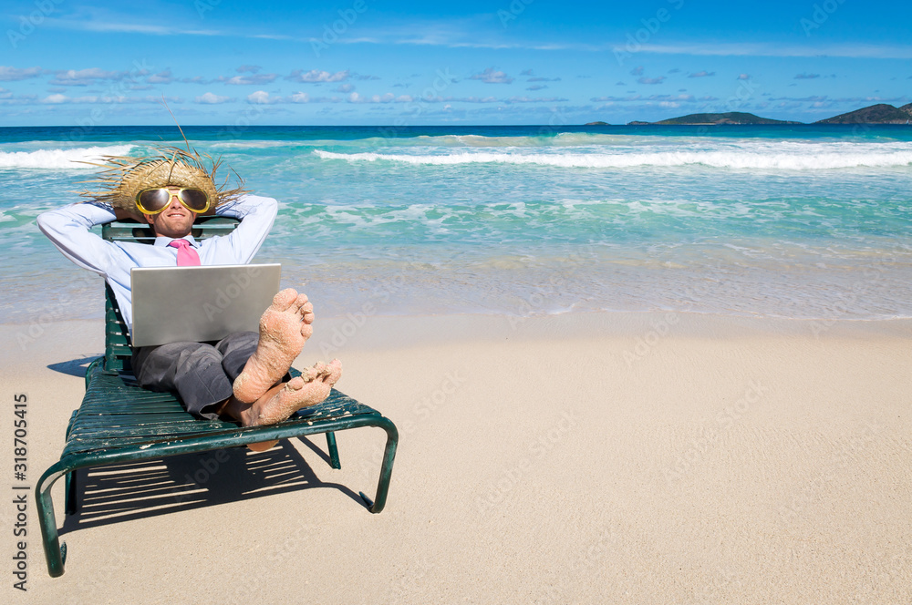 Barefoot businessman kicking back in a beach chair with laptop on his lap and sunglasses and straw hat on his head