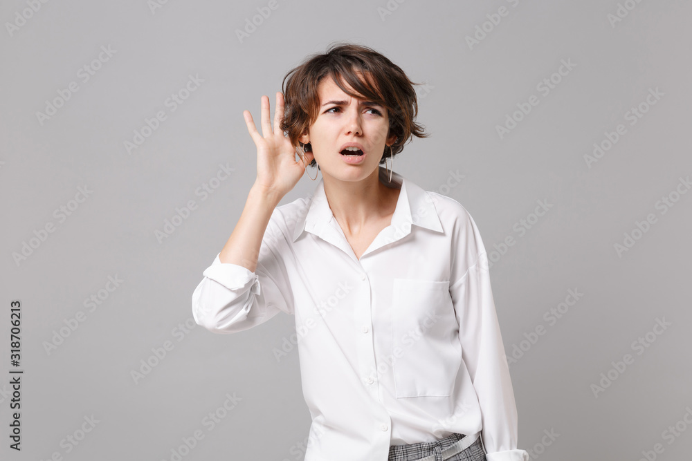 Bewildered young business woman in white shirt posing isolated on grey background studio portrait. Achievement career wealth business concept. Mock up copy space. Try to hear you with hand near ear. Bewildered young business woman in white shirt posing isolated on grey background studio portrait. Achievement career wealth business concept. Mock up copy space. Try to hear you with hand near ear.