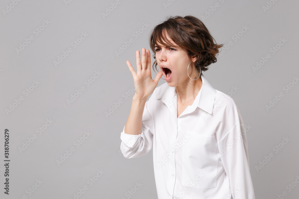 Dissatisfied young business woman in white shirt posing isolated on grey background in studio. Achievement career wealth business concept. Mock up copy space. Screaming with hand gesture near mouth. Dissatisfied young business woman in white shirt posing isolated on grey background in studio. Achievement career wealth business concept. Mock up copy space. Screaming with hand gesture near mouth.