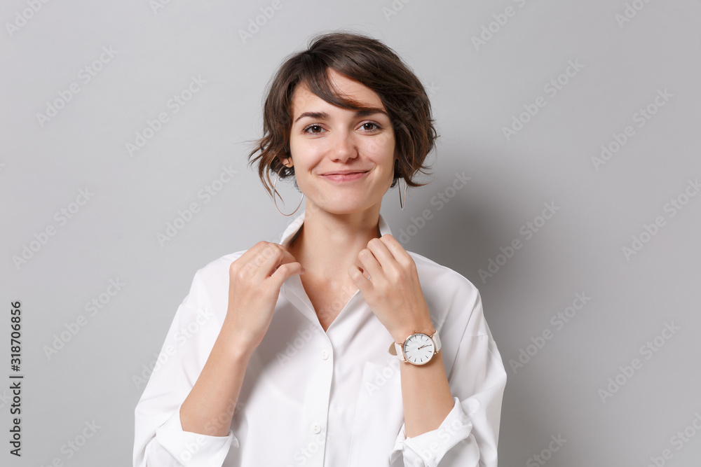 Smiling attractive young business woman in white shirt posing isolated on grey wall background studio portrait. Achievement career wealth business concept. Mock up copy space. Straightening collar.