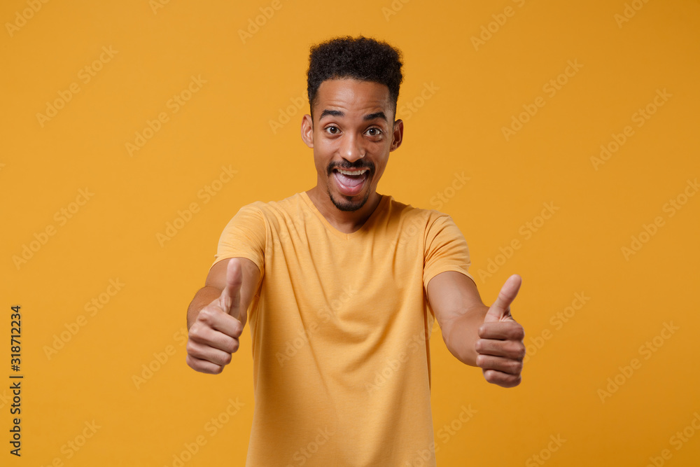 Funny young african american guy in casual t-shirt posing isolated on yellow orange wall background, studio portrait. People sincere emotions lifestyle concept. Mock up copy space. Showing thumbs up. Funny young african american guy in casual t-shirt posing isolated on yellow orange wall background, studio portrait. People sincere emotions lifestyle concept. Mock up copy space. Showing thumbs up.
