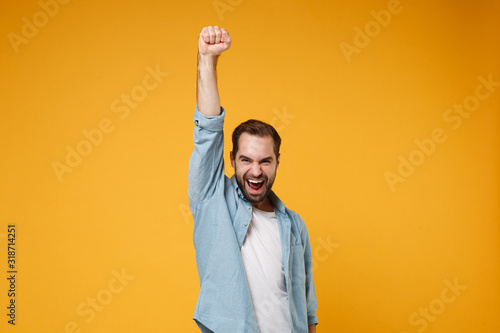 Joyful young bearded man in casual blue shirt posing isolated on yellow orange wall background, studio portrait. People lifestyle concept. Mock up copy space. Clenching fist like winner, rising hand.