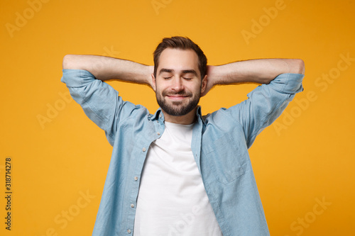 Relaxed young bearded man in casual blue shirt posing isolated on yellow orange background studio portrait. People sincere emotions lifestyle concept. Mock up copy space. Sleep with hands behind head.