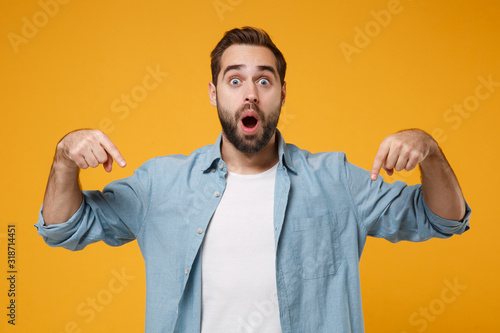 Shocked young bearded man in casual blue shirt posing isolated on yellow orange wall background in studio. People lifestyle concept. Mock up copy space. Pointing index fingers down keeping mouth open. photo