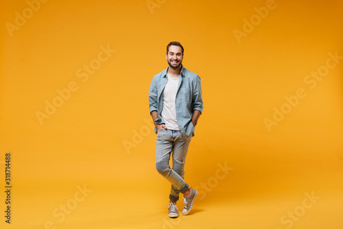 Smiling young bearded man in casual blue shirt posing isolated on yellow orange background, studio portrait. People sincere emotions lifestyle concept. Mock up copy space. Holding hands in pockets.
