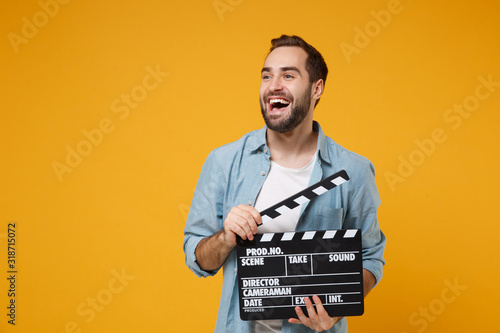 Cheerful young man in casual blue shirt posing isolated on yellow orange wall background, studio portrait. People lifestyle concept. Mock up copy space. Hold classic black film making clapperboard.